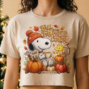 Op de afbeelding: Een beige T-shirt met korte mouwen en een afbeelding van Snoopy met een oranje gebreide muts, een koffiemok vasthoudend, omringd door pompoenen, herfstbladeren en Woodstock. De tekst luidt: Fall is my favorite color, Coffee & Leaves, Cozy Days Ahead en Sweater Weather.