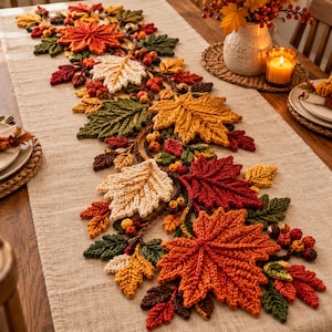 Puede incluir: Un camino de mesa rectangular de color beige decorado con una guirnalda central de hojas de otoño y bayas tejidas a ganchillo. Las hojas presentan tonos naranja, rojo, amarillo, crema y verde oscuro con texturas de punto detalladas.