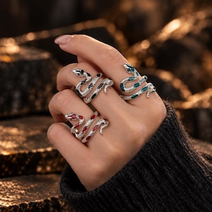 Peut inclure: Trois bagues serpent en métal argenté avec des écailles texturées et des pierres incrustées. Une bague présente des pierres noires, une autre des pierres rouge foncé et la troisième des pierres bleu sarcelle. Chaque bague s'enroule.