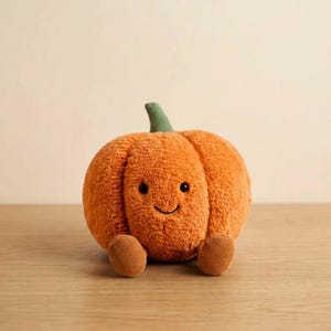 Könnte beinhalten: Ein Kürbis-Plüschtier mit weichem orangefarbenem Fell, einem grünen Stiel und zwei kleinen braunen Füßen aus Cord. Der Kürbis hat ein aufgesticktes lächelndes Gesicht mit zwei schwarzen runden Augen und einem geschwungenen Mund.