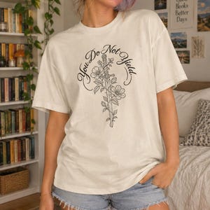 May include: An off-white short-sleeve t-shirt featuring a black line art graphic of a sword entwined with flowers. The text "You Do Not Yield" is printed in a curved, elegant script font above and around the central sword and floral design.