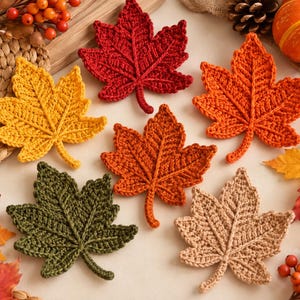 May include: A collection of six crocheted maple leaf appliques in various autumn colors including red, yellow, orange, dark green, and beige. Each leaf features detailed stitched veins and a textured surface, arranged in a scattered pattern.