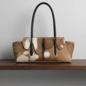Könnte beinhalten: Eine strukturierte Handtasche mit einem Rindsleder-Außenmaterial in einem braun, weiß und schwarz gefleckten Muster. Die Tasche hat eine horizontale rechteckige Form mit Klappverschluss und zwei hohen, dünnen schwarzen Lederhenkeln.