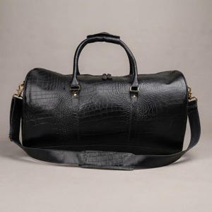 Peut inclure: Un sac de voyage noir avec un motif texturé en peau de crocodile. Le sac comprend deux poignées supérieures, une bandoulière amovible avec une section rembourrée et des attaches en métal doré sur les côtés pour fixer la bandoulière.