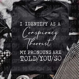 May include: A black t-shirt featuring white text that reads I IDENTIFY AS A Conspiracy Theorist MY PRONOUNS ARE TOLD/YOU/SO. The shirt is displayed alongside a black bag with a white spiderweb design and black and white chequered slip-on shoes.