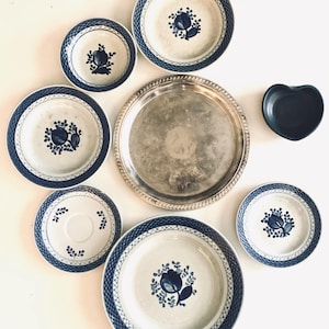 May include: A collection of ceramic tableware featuring a white base with blue floral patterns and lattice borders, including five round plates of varying sizes, one large circular silver-toned metal tray, and one small dark gray heart-shaped bowl.