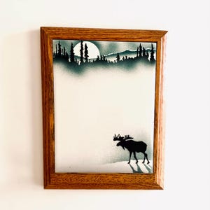 May include: A rectangular wall art piece featuring a silhouette of a moose standing on a snowy landscape, with a dark forest and a large full moon in the background. The artwork is encased in a medium-toned wooden frame with a visible grain.