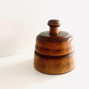 May include: A round, medium-brown wooden butter stamp featuring a smooth, tiered cylindrical body and a small, rounded knob handle on top. The wood grain is visible throughout the surface, showing natural variations in color and texture.