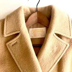 May include: Close-up of a tan wool coat with a notched collar and a button closure. The coat is hanging on a wooden hanger. A label inside the coat reads "PENDLETON WOOLEN MILLS" and "MADE IN USA".