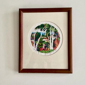 May include: Framed artwork featuring a circular embroidered landscape scene. The artwork depicts trees, buildings, and colorful foliage. The frame is a dark brown wood. Text on the artwork reads "Beauty's view" and "Jacqueline Fulker 1997".