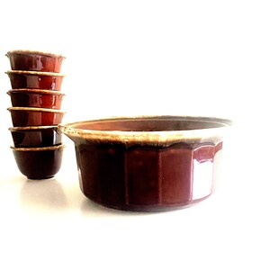 May include: A collection of brown ceramic bowls, including a large serving bowl and six smaller bowls stacked on the left. The bowls have a glossy finish and a contrasting light-colored rim. The serving bowl has a faceted design.