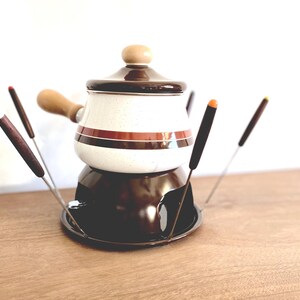 May include: A white ceramic fondue pot with brown and orange stripes, a brown lid, and a brown metal base. The pot is surrounded by six fondue forks with brown wooden handles and orange tips.