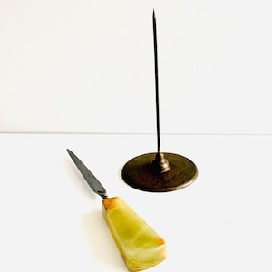 May include: A metal paper spike with a circular base and a letter opener featuring a tapered, light green, translucent stone handle with brown veining and a pointed, dark metal blade, both items displayed together on a plain white surface.
