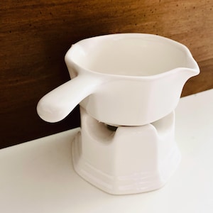 May include: A white ceramic fondue pot with a single handle and a pouring spout, resting on a matching white ceramic base. The pot features a smooth, glossy finish and a rounded bowl shape, while the base has a sturdy, open-frame design.