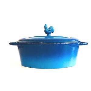May include: A blue enameled cast iron Dutch oven with a matching lid. The lid features a decorative rooster handle. The oven has two side handles and a gradient color from a darker blue to a lighter blue.