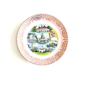 May include: A vintage decorative plate featuring a pink border with gold filigree and a central image of Washington D.C. landmarks. The plate includes images of the Capitol Building, the White House, and the Washington Monument.