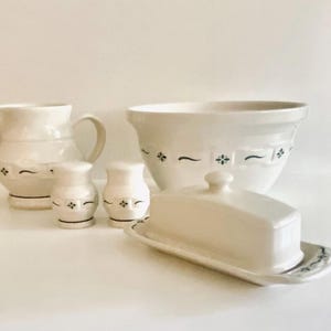 May include: A collection of white ceramic kitchenware with a green floral and line design. Includes a pitcher, mixing bowl, butter dish, and two salt and pepper shakers. The items are arranged on a white surface.