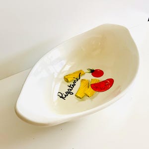 May include: White ceramic serving dish with a curved, oblong shape. The dish is decorated with a Rigatoni pasta illustration, including yellow pasta, a red tomato, and the word "Rigatoni" in black script.