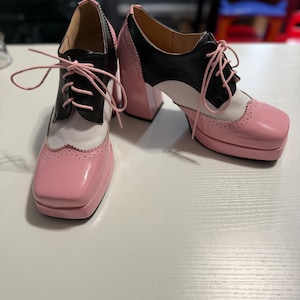 May include: A pair of lace-up platform heels featuring a color-blocked design in glossy pink, black, and white. These shoes have a square toe, a thick block heel, and decorative brogue-style perforations along the edges of the pink toe cap.
