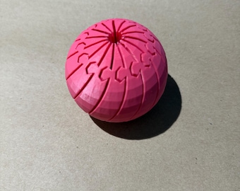 Puede incluir: Un objeto esférico de color rosa brillante que presenta una serie de ranuras radiales y patrones geométricos entrelazados que convergen en una abertura circular central en la superficie superior, creando una apariencia texturizada y simétrica.