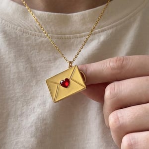 Puede incluir: Un collar de tono dorado con un colgante rectangular en forma de sobre. El colgante presenta una pequeña piedra roja vibrante en forma de corazón centrada en la solapa. La pieza cuelga de una delicada cadena dorada sobre una camiseta color crema.
