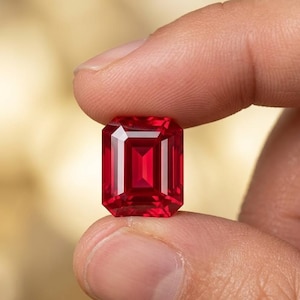 May include: A vibrant, deep red gemstone cut in a rectangular emerald shape with faceted edges. The stone displays high clarity and a rich, saturated crimson hue, reflecting light through its geometric facets to create a brilliant, polished appearance.