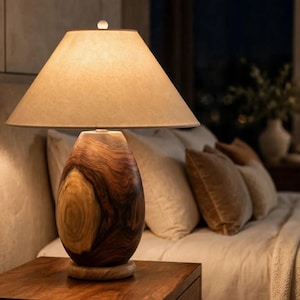 May include: A table lamp featuring a smooth, oval-shaped wooden base with prominent dark brown and light tan wood grain patterns. The lamp is topped with a wide, conical, off-white fabric lampshade that emits a soft, warm glow from within.
