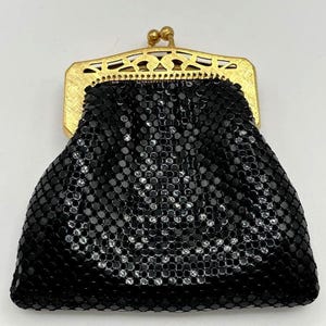 May include: A black mesh coin purse featuring a gold-toned metal frame with a decorative cutout pattern and a dual ball clasp closure. The body of the purse is constructed from small, interlocking black metal links that create a flexible texture.
