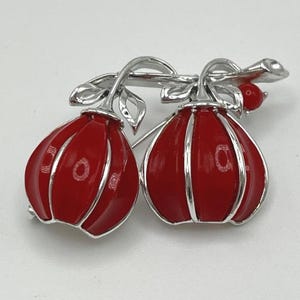 May include: A silver-toned brooch featuring two rounded, red enameled fruit shapes hanging from a silver stem with leaves. The design includes vertical silver ridges on each red fruit and a small, round red bead accent near the top right stem.