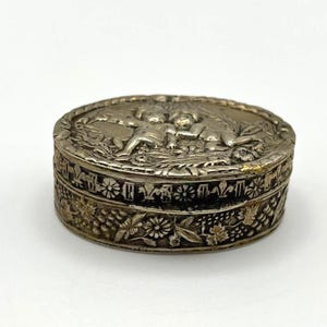 May include: An oval silver-toned metal trinket box featuring an embossed lid with two cherubs surrounded by foliage. The sides of the box are decorated with a repeating pattern of flowers, leaves, and grape clusters in a dark, oxidized finish.