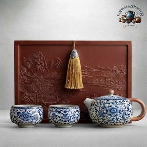 May include: A tea set featuring a blue and white floral patterned teapot with a wooden handle and two matching tea cups. Behind the set is a rectangular brown clay tray with an embossed landscape scene and a hanging gold tassel.