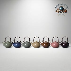 Puede incluir: Siete teteras de cerámica dispuestas en fila, cada una con un cuerpo redondeado, una tapa pequeña, un pico curvo y un asa arqueada oscura. Las teteras tienen acabados en colores sólidos que incluyen verde salvia, azul, gris, beige, marrón, rosa y morado oscuro.