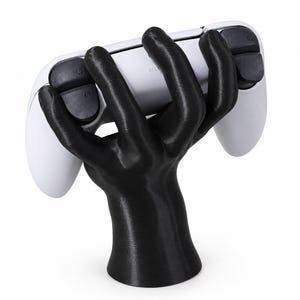 May include: A black hand-shaped stand holding a white video game controller. The controller features black L1, L2, R1, and R2 shoulder buttons. The stand is sculpted in a grasping position, providing a stable base for the gaming device.