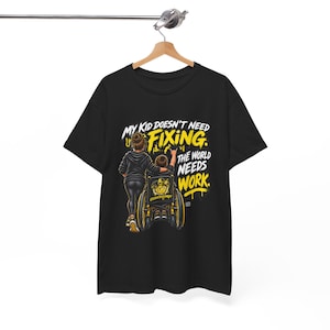 Puede incluir: Una camiseta negra de manga corta con una ilustración gráfica de una persona de pie detrás de un niño en silla de ruedas. El texto dice: MY KID DOESN'T NEED FIXING. THE WORLD NEEDS WORK. El diseño utiliza letras y detalles en amarillo y blanco.