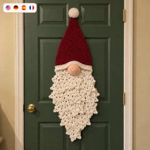 May include: A crochet gnome wall hanging featuring a tall, pointed dark red hat topped with a white pom-pom, a round peach-colored nose, and a long, textured white beard made of intricate, layered crochet stitches hanging vertically.