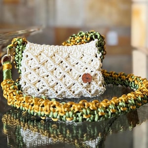 May include: A small macrame handbag featuring an off-white diamond-patterned body with a thick, braided shoulder strap in alternating shades of olive green and mustard yellow. A circular brown leather tag with a stamped logo is attached to the front.