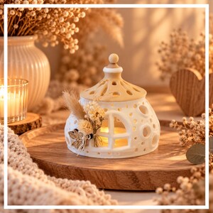 Puede incluir: Un portavelas de cerámica blanca con forma de casita y techo color canela, con aberturas circulares y una ventana. Un pequeño ramo de flores secas y un dije de hoja de madera están sujetos al lateral, con una luz cálida brillando en su interior.