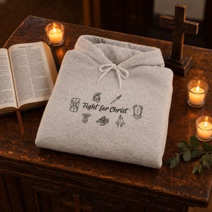 Puede incluir: Una sudadera con capucha de color gris claro que presenta un estampado gráfico negro con el texto "Fight for Christ" rodeado de ilustraciones de un escudo, casco, espada, coraza, sandalias, cinturón y manos en oración con cordones blancos.