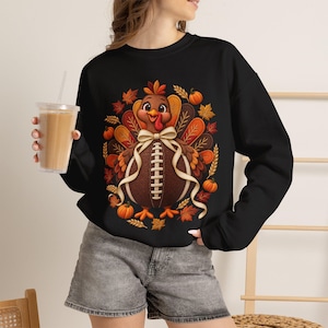 Op de afbeelding: Een zwart sweatshirt met lange mouwen en een afbeelding van een kalkoen met een lichaam dat lijkt op een football, voorzien van een crèmekleurige strik. Het ontwerp is omringd door herfstachtige elementen zoals oranje pompoenen, korenaren en kleurrijke herfstbladeren.