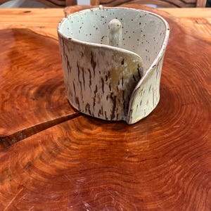 May include: A cylindrical ceramic ring holder with an overlapping edge design. The exterior features a textured, mottled tan and brown finish, while the interior is off-white with dark speckles and a central vertical post for rings.