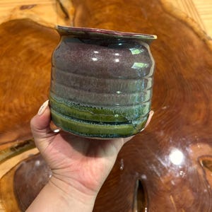 Peut inclure: Un petit mug en céramique avec un vernis brillant, présentant une section supérieure violet foncé et une section inférieure vert vif. Le mug possède un corps arrondi et nervuré avec des indentations horizontales et un bord évasé.
