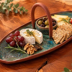 May include: An oval-shaped ceramic serving tray with a terracotta-coloured handle and a teal-glazed surface featuring a textured lace pattern. The tray holds a small bowl of olives, a wheel of brie cheese, red grapes, walnuts, crackers, and tomatoes.