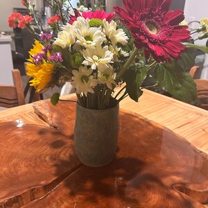 May include: A bouquet of flowers in a speckled grey ceramic vase. The arrangement includes a large dark red gerbera daisy, white daisies with green centres, yellow sunflowers, orange carnations, and small purple filler flowers with green stems.