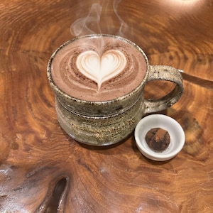 Peut inclure: Une tasse en céramique mouchetée remplie d'une boisson au chocolat chaud fumante avec un motif de latte art en forme de cœur blanc à la surface, accompagnée d'un petit bol blanc contenant une truffe au chocolat ronde saupoudrée de cacao.