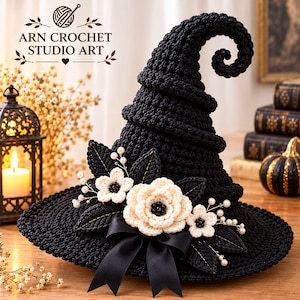 Pode incluir: Um chapéu de bruxa preto em crochê com ponta curvada, adornado com flores de crochê na cor creme, folhas escuras, pequenas contas brancas tipo pérola e um laço de fita de cetim preta. O texto ARN CROCHET STUDIO ART é visível no canto superior esquerdo.