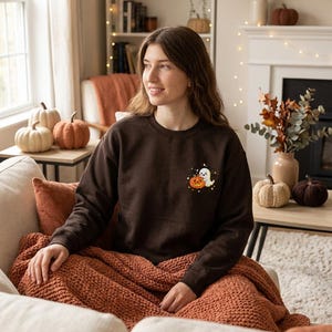 Könnte beinhalten: Ein dunkelbraunes Sweatshirt mit Rundhalsausschnitt und einer kleinen Stickerei auf der linken Brust. Das Motiv zeigt ein weißes Gespenst, das einen lächelnden orangefarbenen Kürbis hält, umgeben von kleinen gelben Sternen.