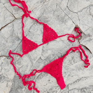 May include: A bright red, handmade crochet bikini set. The top is a triangle style, and the bottom is a thong. The bikini is displayed on a gray, textured rock surface. The crochet work has an open, lacy pattern.
