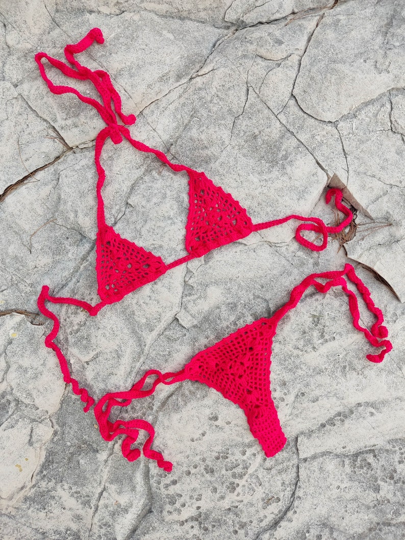 May include: A bright red, handmade crochet bikini set. The top is a triangle style, and the bottom is a thong. The bikini is displayed on a gray, textured rock surface. The crochet work has an open, lacy pattern.