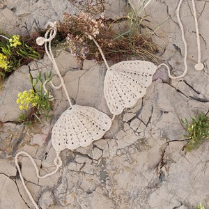 Puede incluir: Un top de bikini de crochet de color crema, sobre una superficie de roca gris claro agrietada. El top está hecho de un tejido delicado y de tejido abierto con un borde festoneado. El top está atado con cuerdas finas.