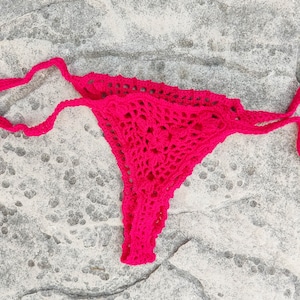 May include: A bright pink, handmade crochet bikini bottom. The bottom features a delicate, open weave design and string ties. The garment is displayed on a textured, gray stone surface. The crochet work appears to be of a medium weight yarn.